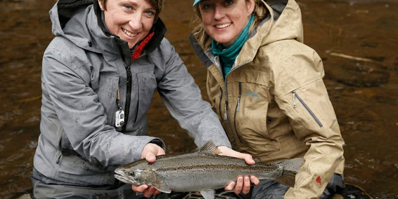 Two women anglers holding a fish in a river while wearing cold-weather fishing jackets
