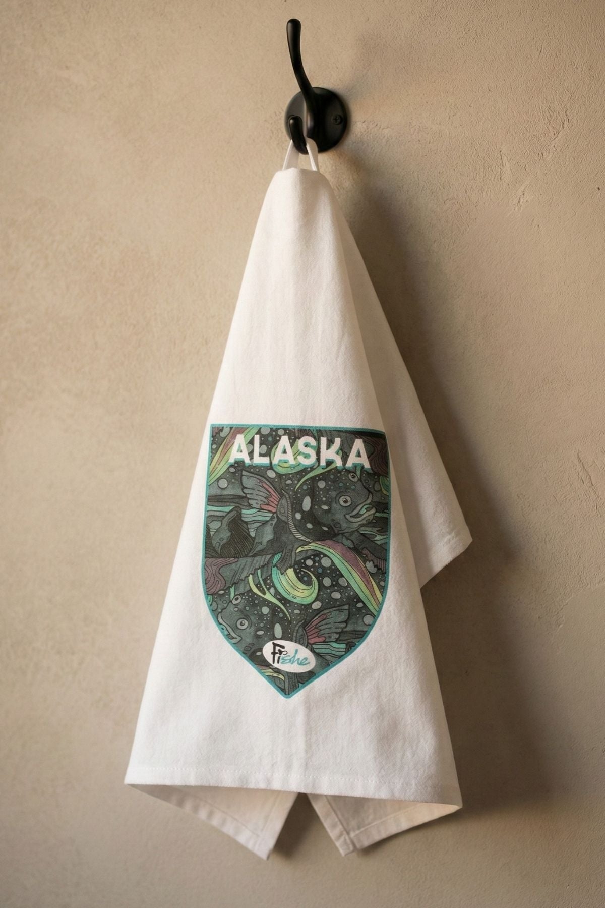White towel with Alaska graphic hanging on a hook against a beige wall