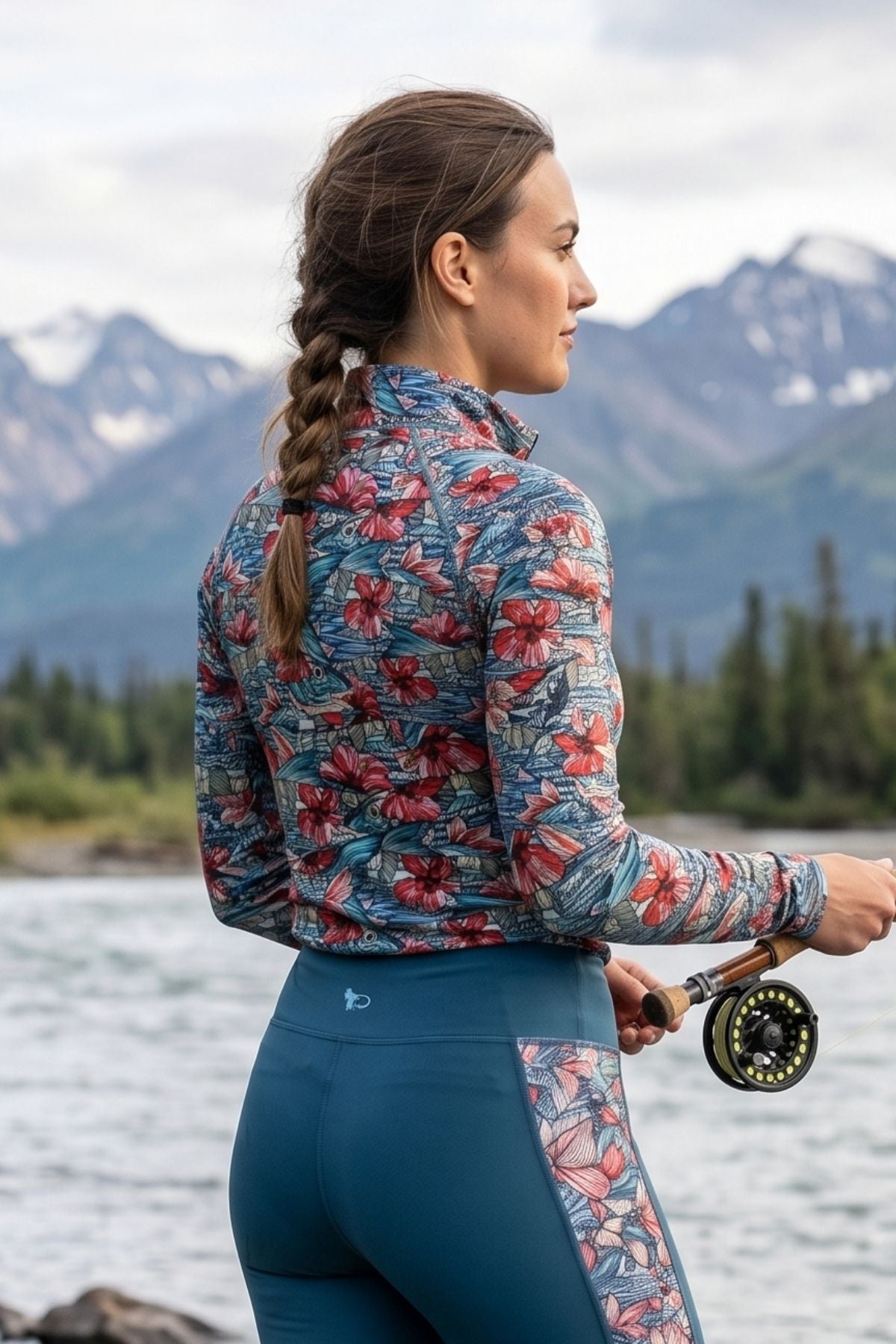 Woman in a colorful jacket and blue pants fishing by a lake with mountains in the background