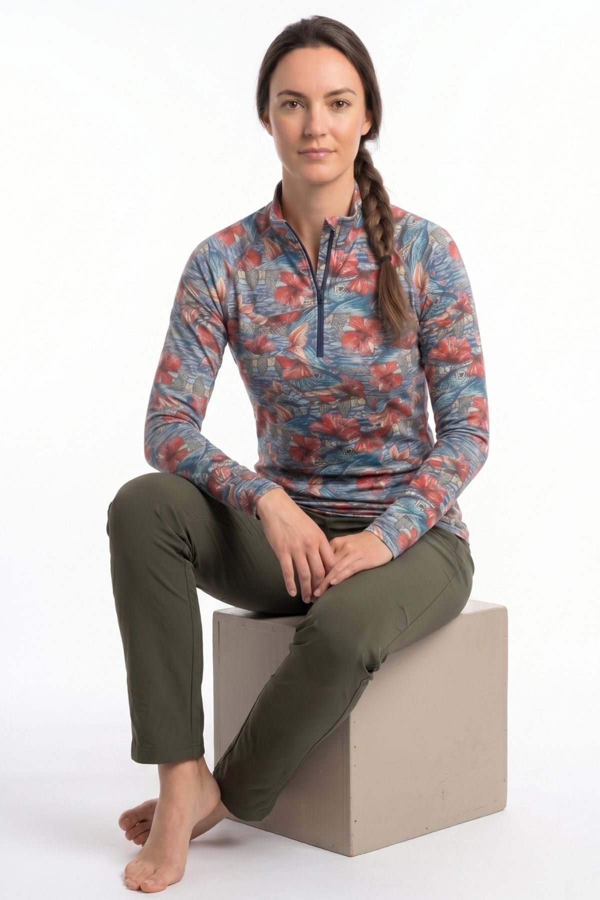 Woman wearing a colorful floral long-sleeve shirt and green pants sitting on a white cube.