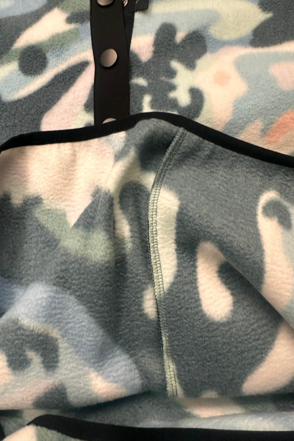 Close-up of a soft fleece blanket with a muted camouflage pattern in gray, blue, beige, and black, featuring black trim and a black strap with silver snaps.