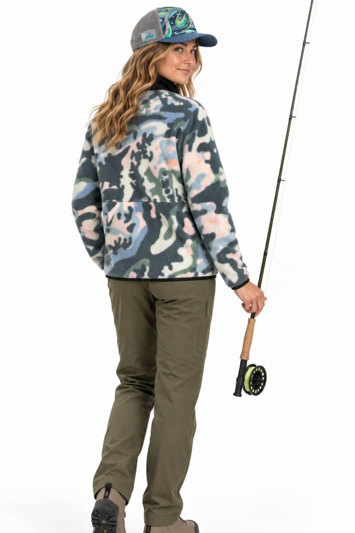 A woman wearing a camouflage fleece jacket, olive green pants, and a patterned cap, holding a fly fishing rod against a white background.