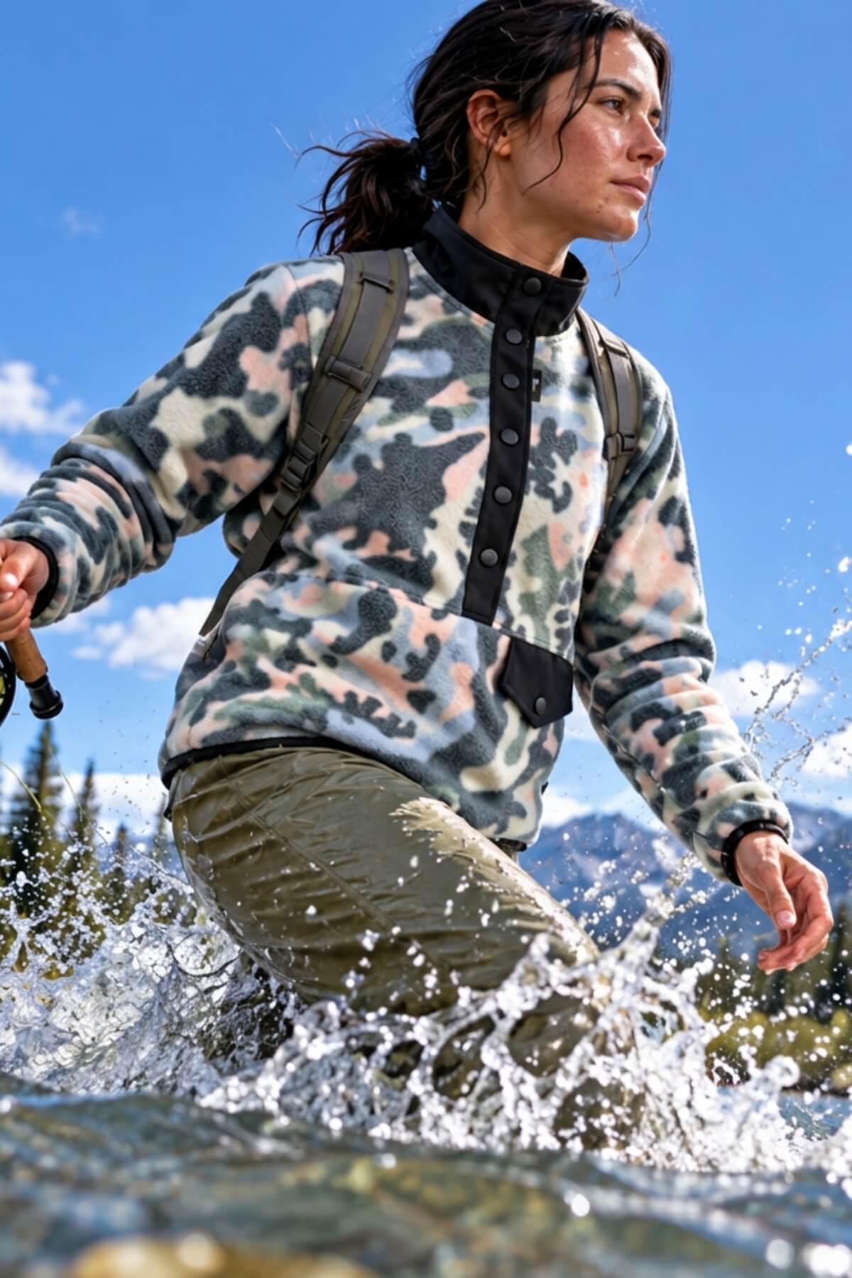 A woman wading through a river wearing a fleece pullover with a green, beige, and pink camouflage pattern, olive pants, and a backpack, with mountains and a blue sky in the background.