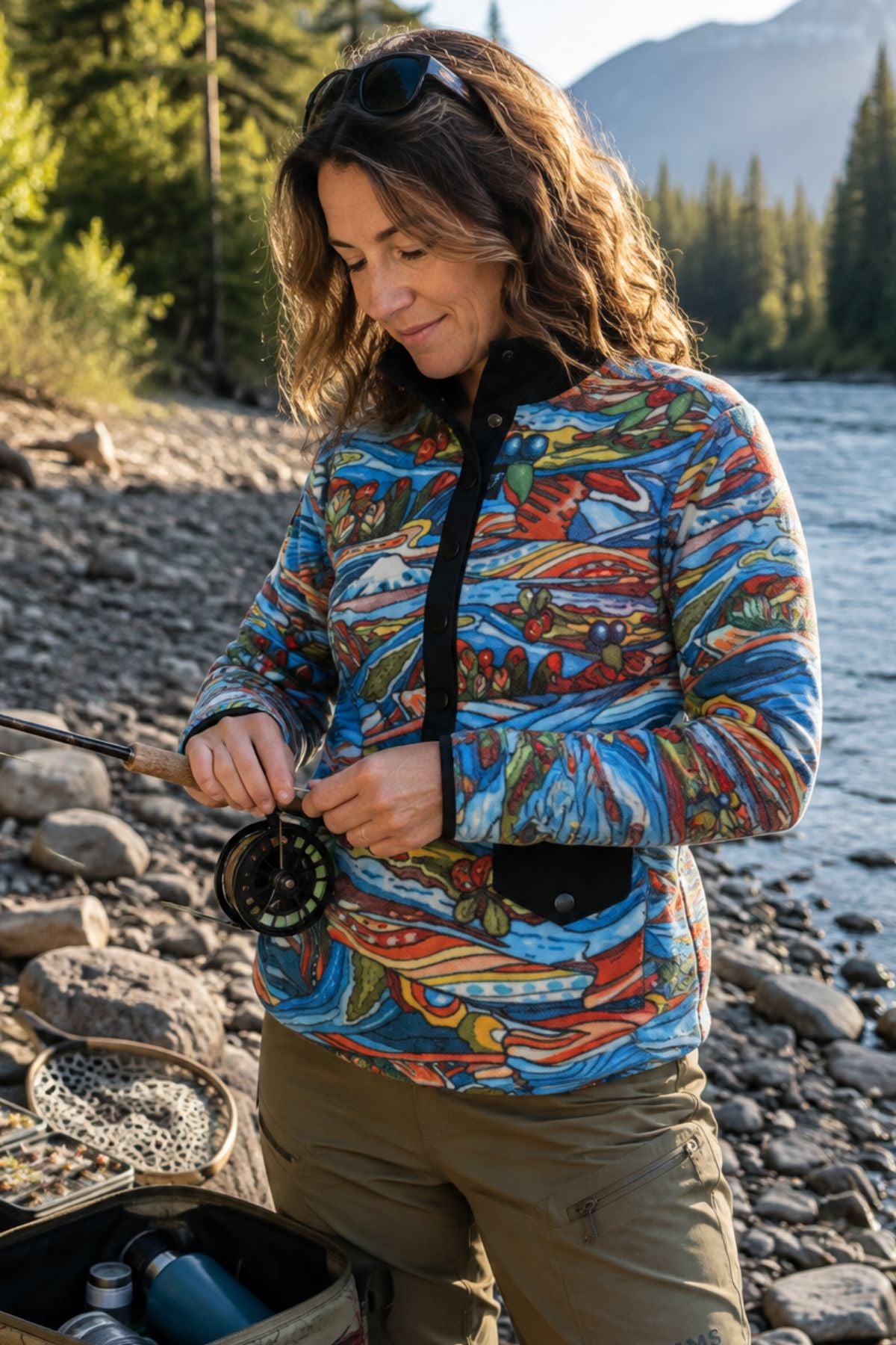 Woman wearing the Charvana Riffle Snap Fleece while preparing fly fishing gear beside a mountain river.