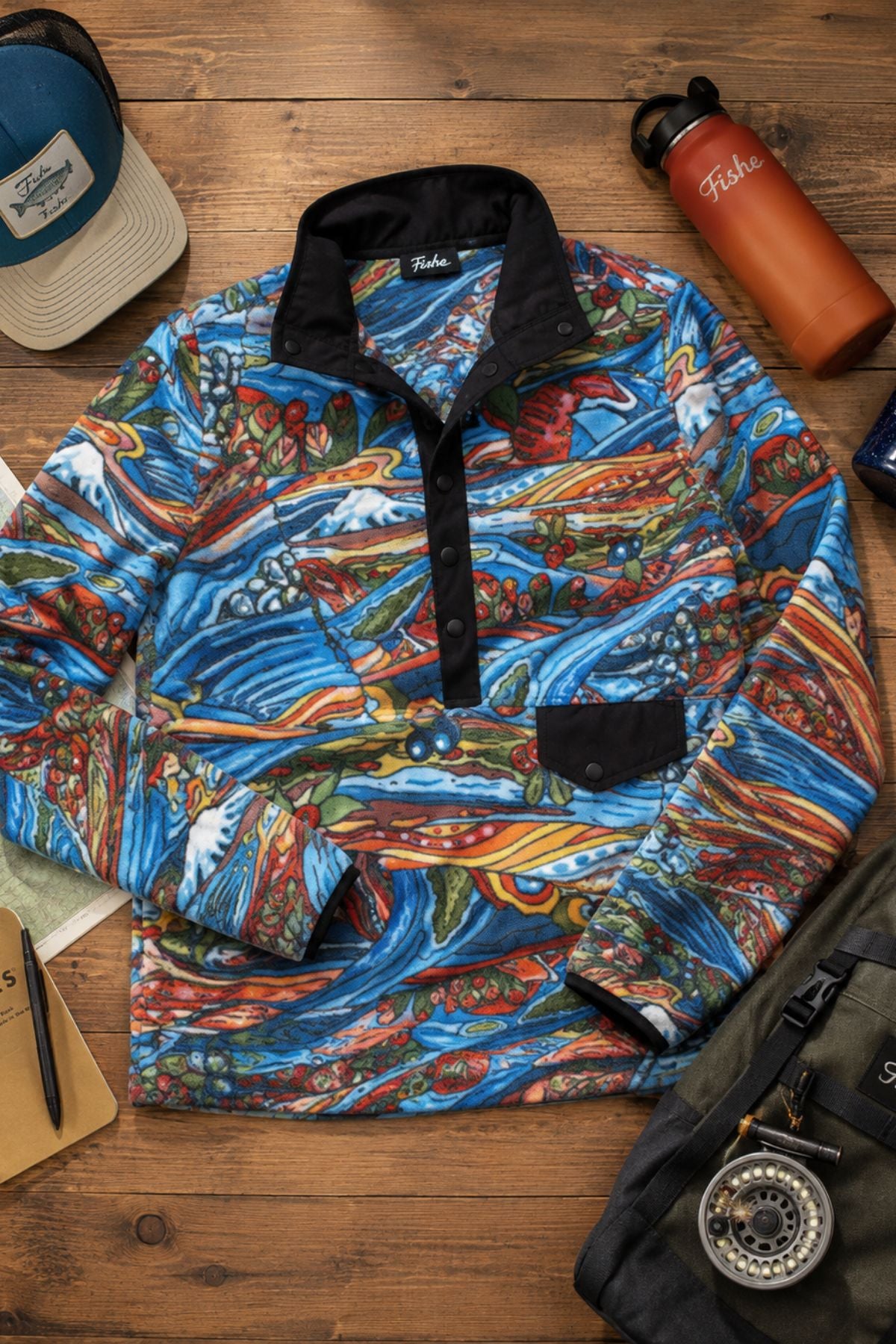 Flat lay of the Charvana Riffle Snap Fleece with fly fishing and outdoor accessories on a wood surface.