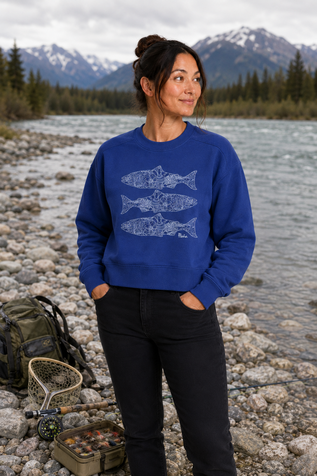 A woman stands on a rocky riverbank wearing a royal blue crewneck sweatshirt with a white fish graphic, paired with black pants. Fishing gear, including a backpack, rod, and tackle box, rests on the rocks beside her, with a river and snow-capped mountains in the background.