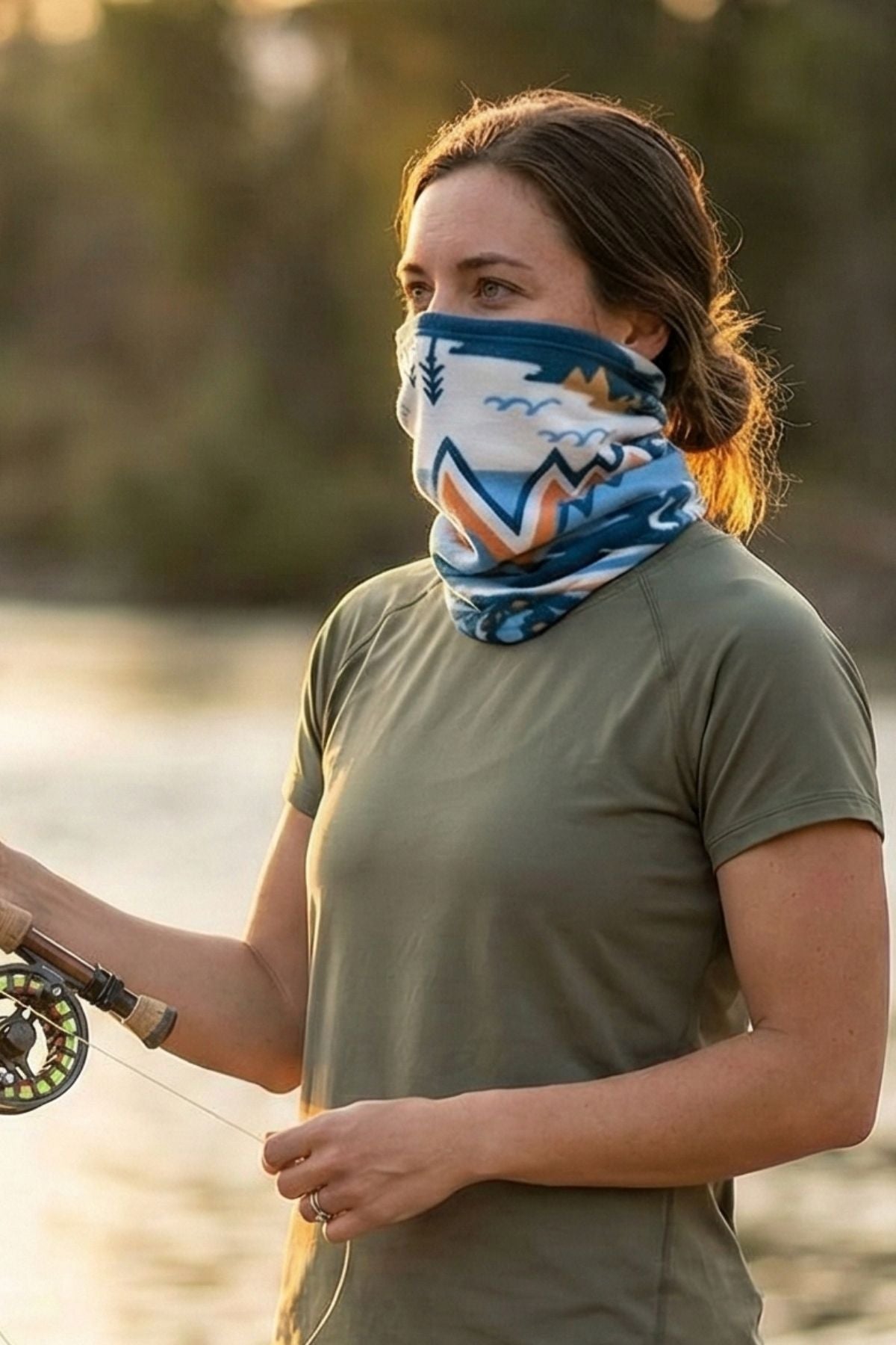 Woman fishing by a body of water wearing a patterned face covering.