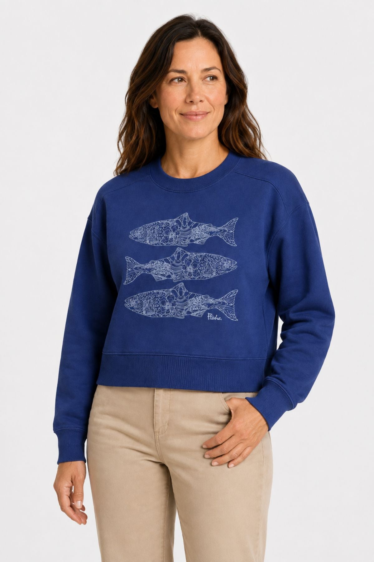 A woman wearing a royal blue cropped crewneck sweatshirt with three white fish illustrations on the front, paired with beige pants, against a light gray studio background.