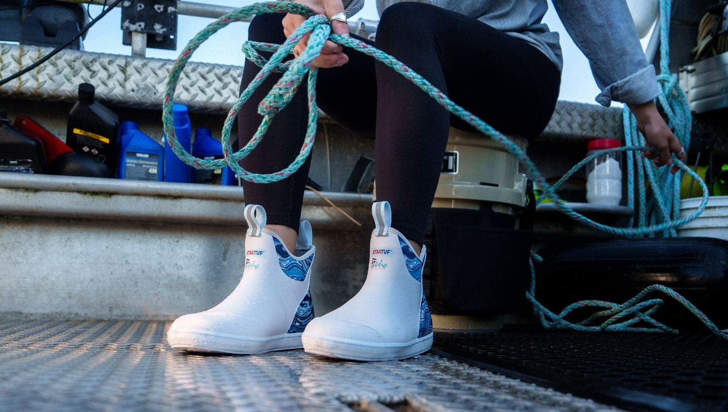 Person wearing white boots with blue patterns, sitting on a truck bed holding green rope.