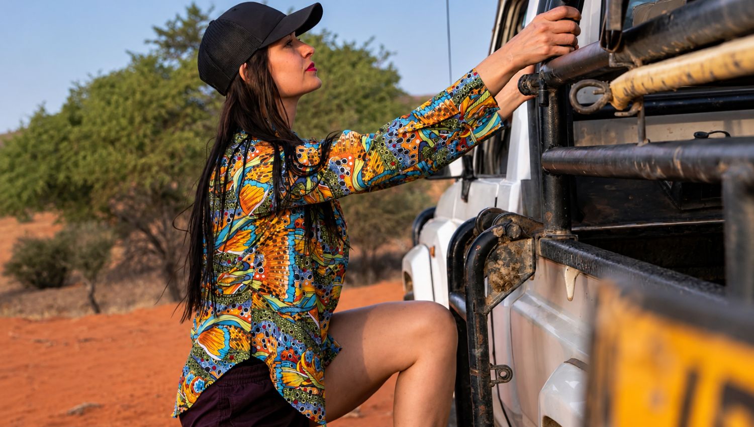 Woman in colorful shirt and black cap interacting with a vehicle in a natural setting
