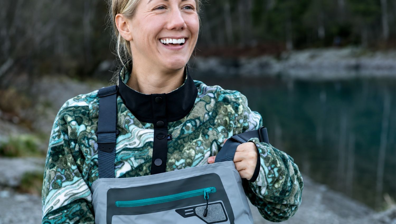 Woman holding a gray bag with a logo, standing outdoors near a body of water.