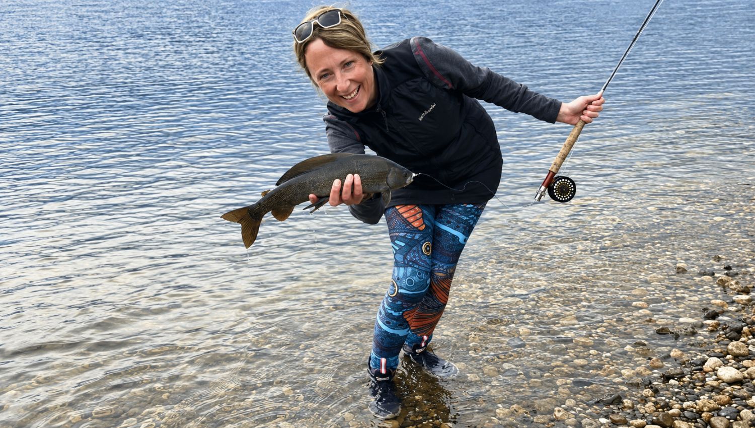 Person holding a fish and a fishing rod by a body of water