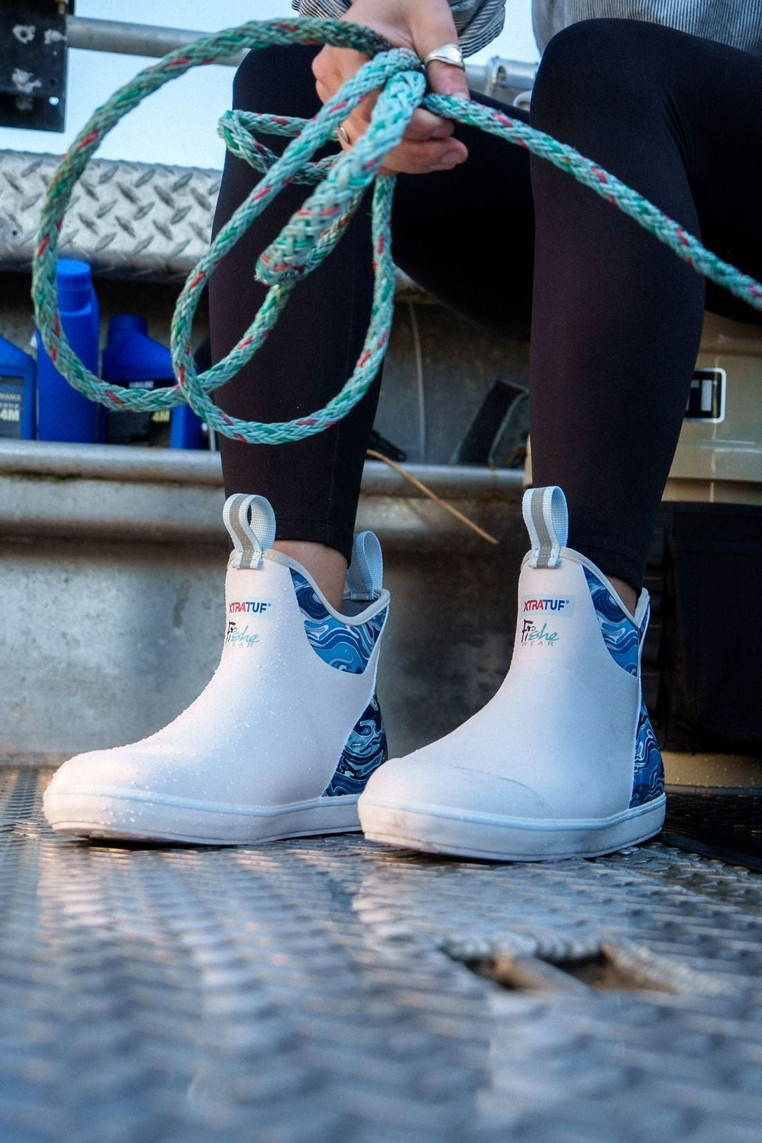 Person wearing white boots with blue patterns, sitting on a metal surface holding green rope.