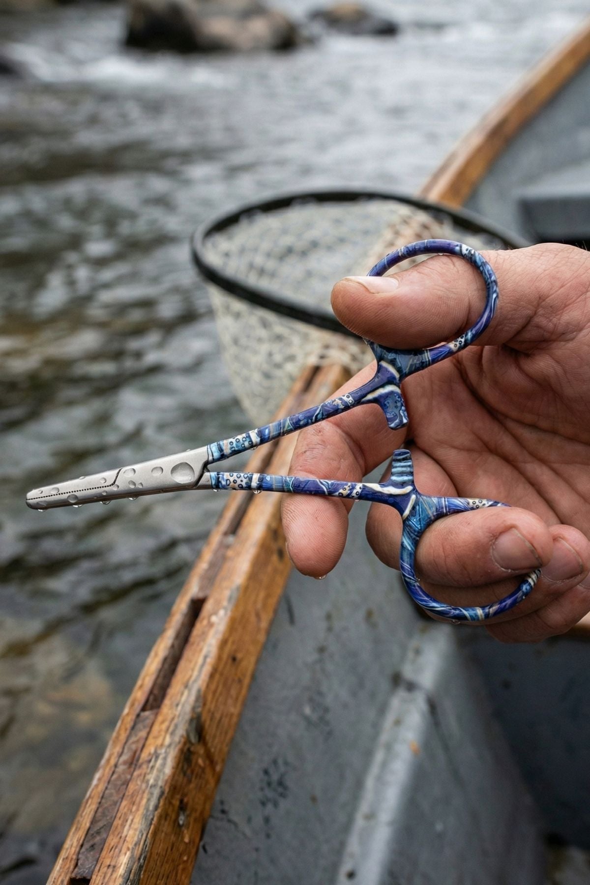 Hand holding a pair of blue-handled scissors with a river and wooden boat in the background