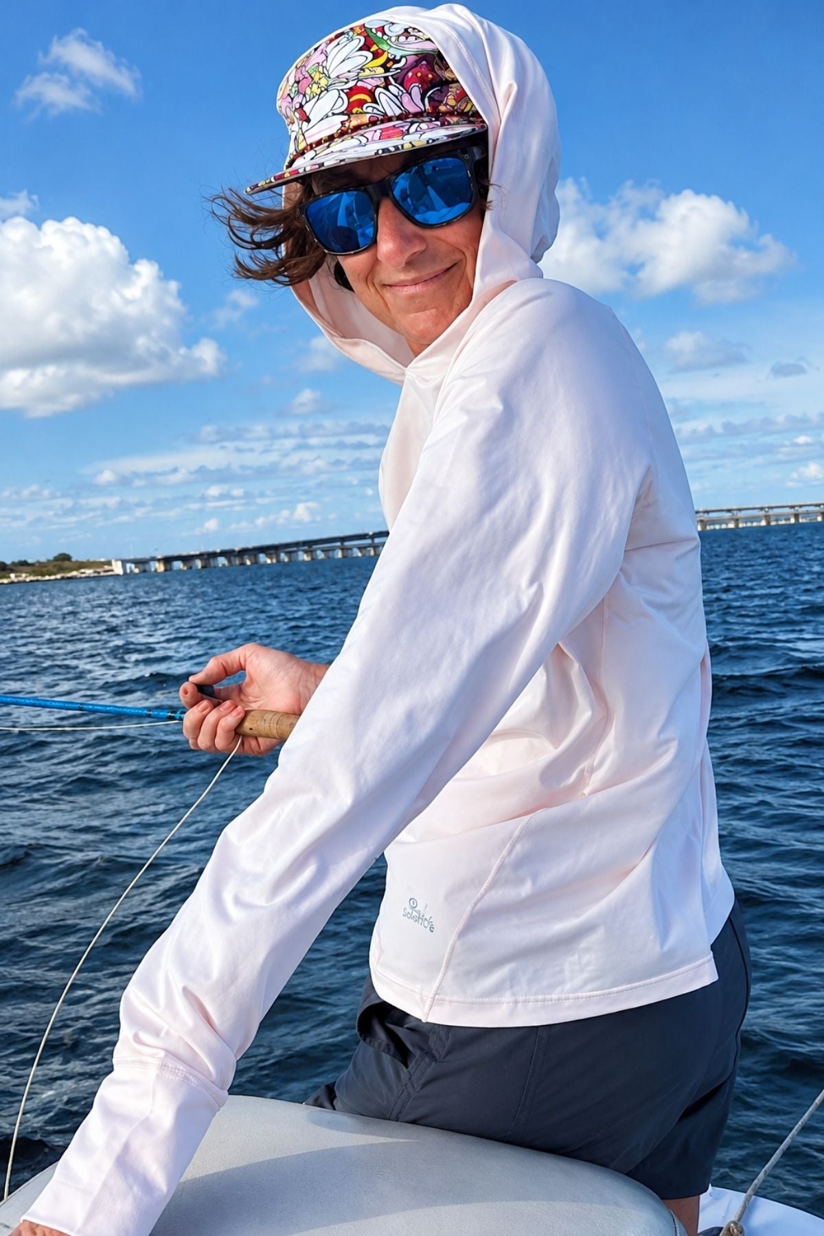 Person on a boat wearing a white hoodie, colorful cap, and sunglasses with a bridge and water in the background.