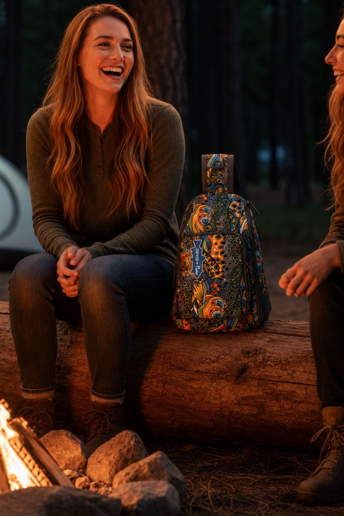 Two women sitting by a campfire with a colorful lantern between them, surrounded by nature.