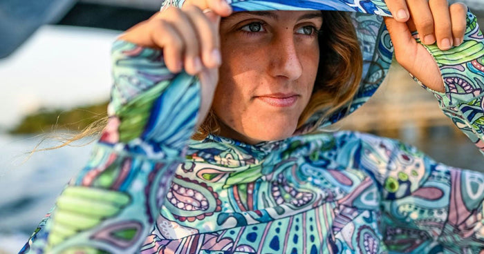 Close-up of an angler adjusting the hood on a Fishe Radiant Redfish Solstice Hooded Sunshirt—bold teal and pink pattern with thumbhole cuffs in sunrise light.