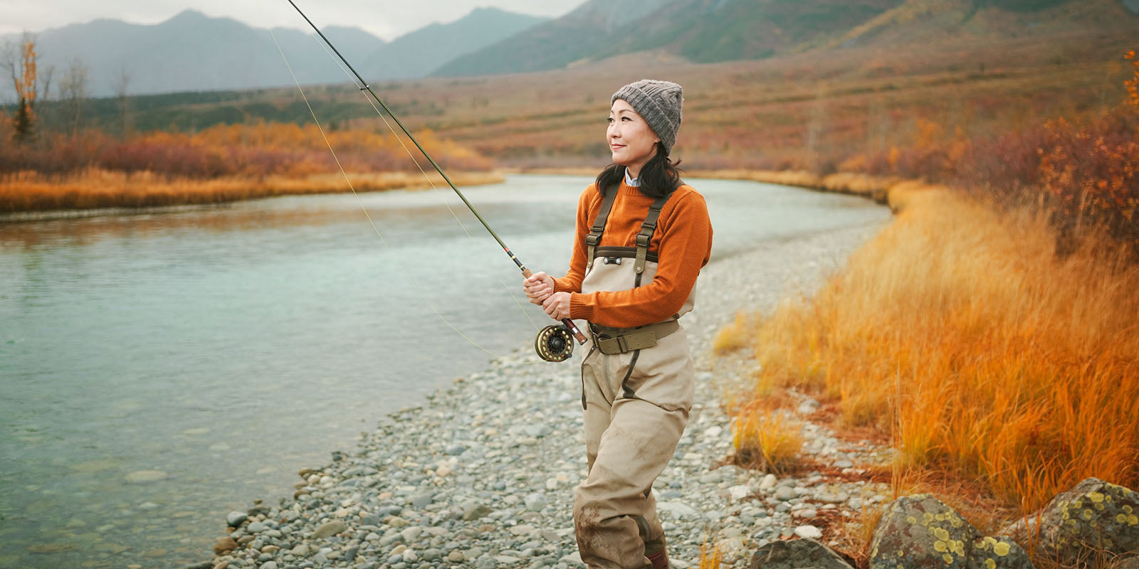 Woman angler in an orange sweater beside an Alaska river in autumn