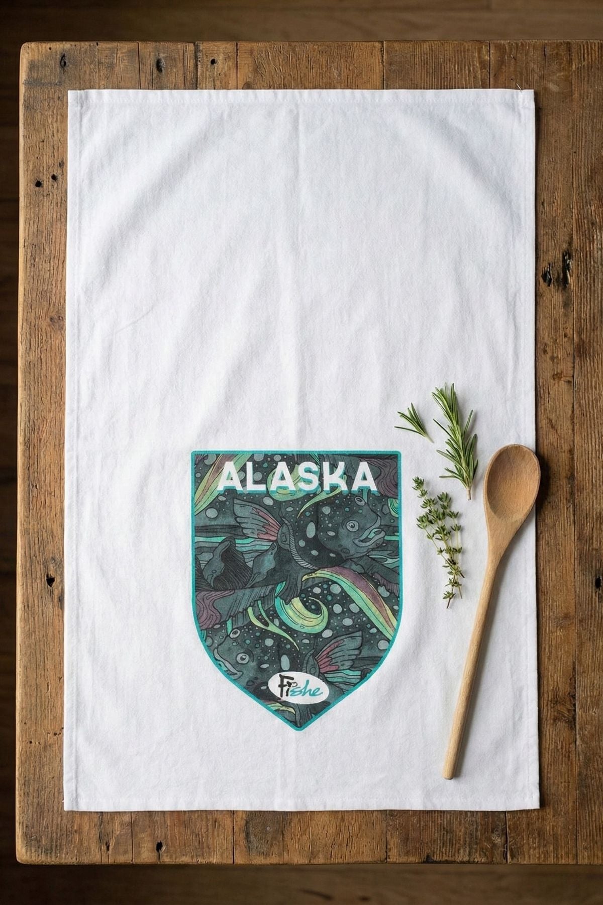 Tea towel with Alaska design on a wooden surface with a wooden spoon and herbs.