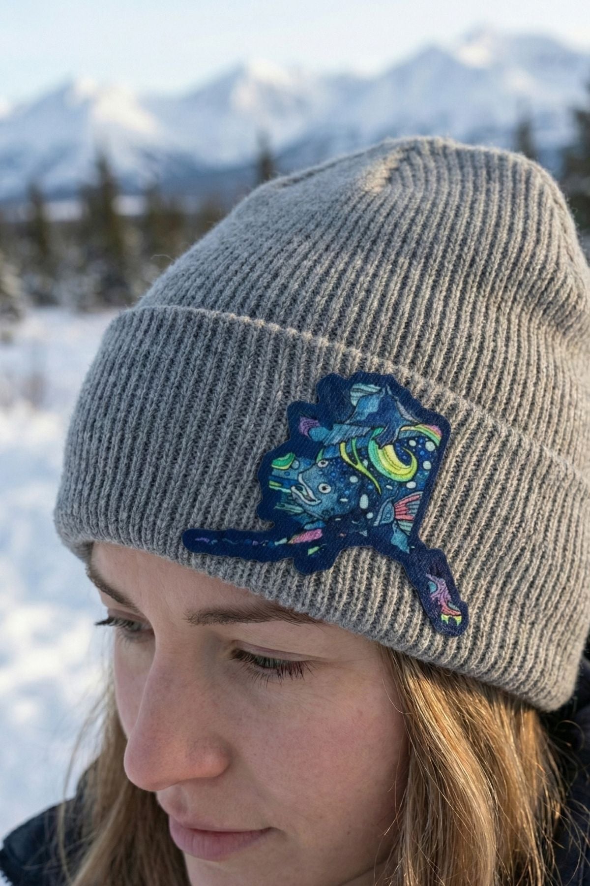 Person wearing a gray beanie with a colorful cat patch in a snowy landscape