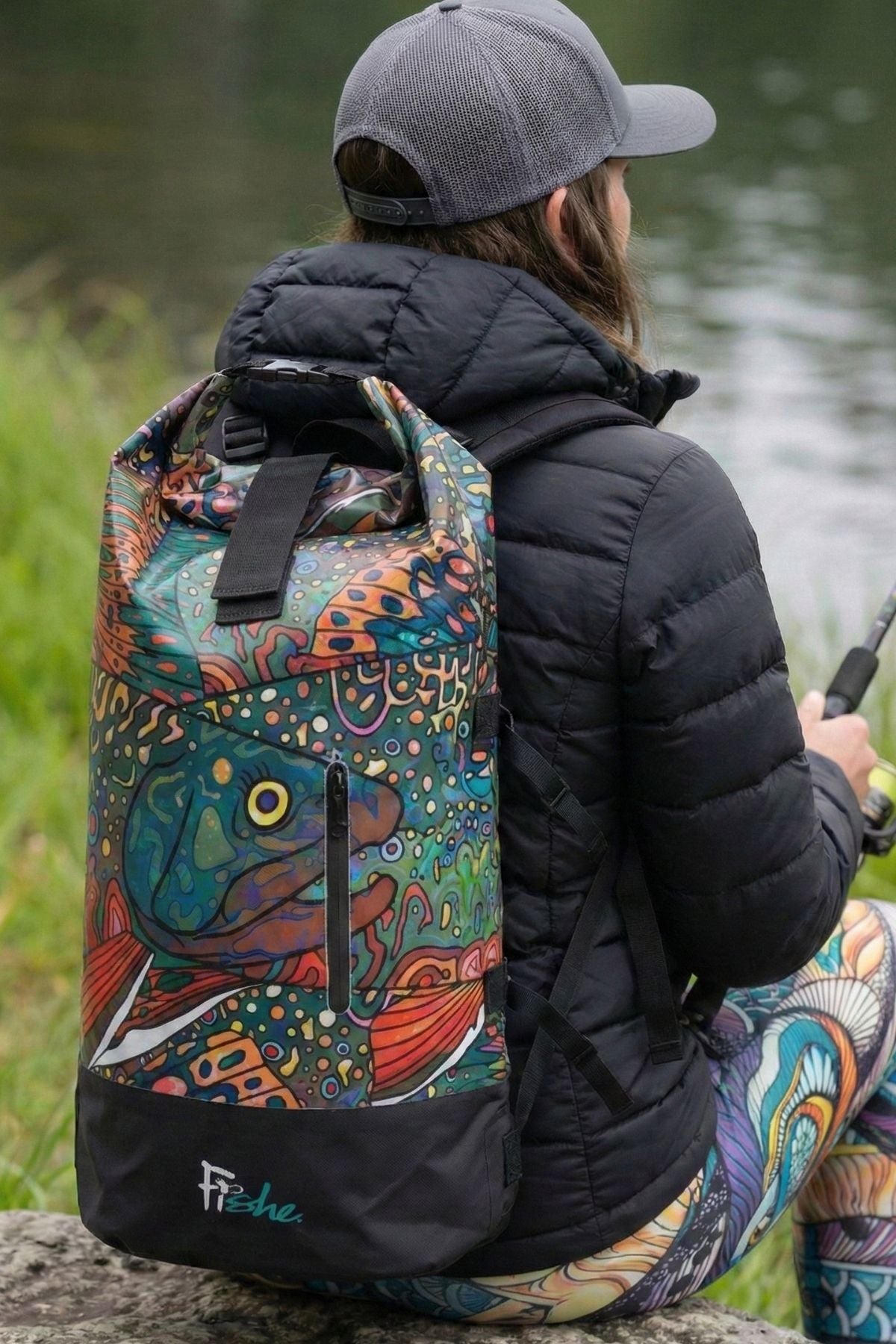 Person with a colorful backpack featuring fish designs by a body of water.
