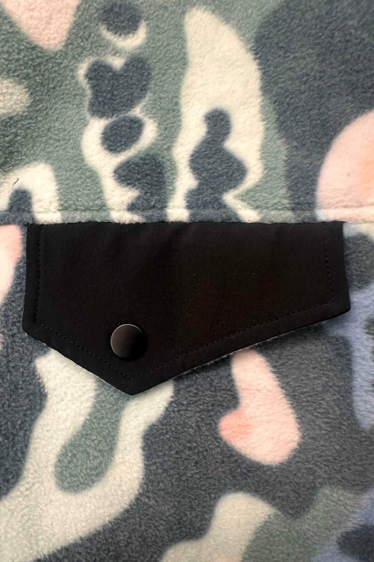 Close-up of a black fabric flap pocket with a snap button closure, laid flat on a soft camouflage-patterned fleece background.