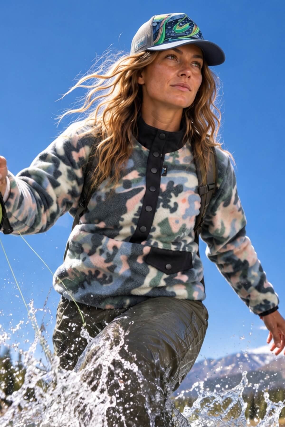 A woman outdoors in a mountain setting wearing a fleece pullover with a muted camouflage pattern in gray, green, and beige, paired with dark pants and a cap, with water splashing around her legs.