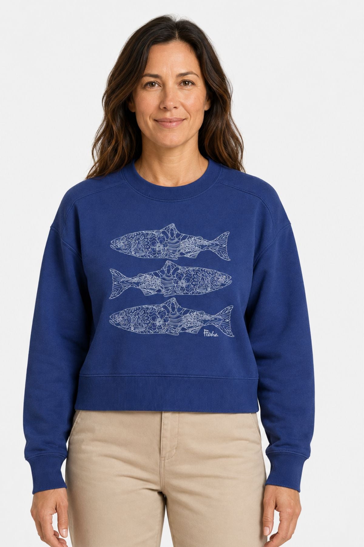 A woman wearing a royal blue cropped sweatshirt with three white fish illustrations on the front, paired with beige pants, against a plain white background.