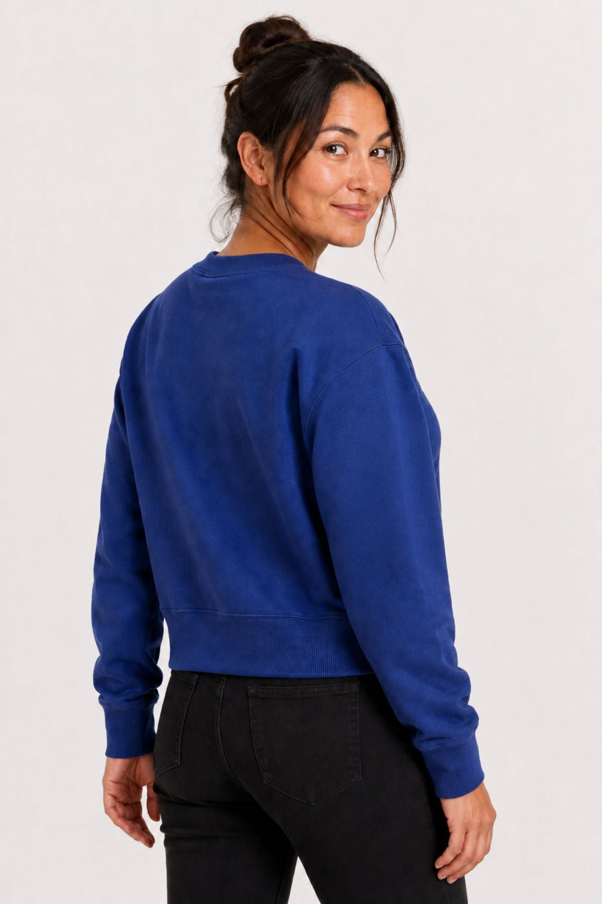 A woman wearing a royal blue cropped crewneck sweatshirt and black pants, shown from a three-quarter back view against a plain light gray studio background.