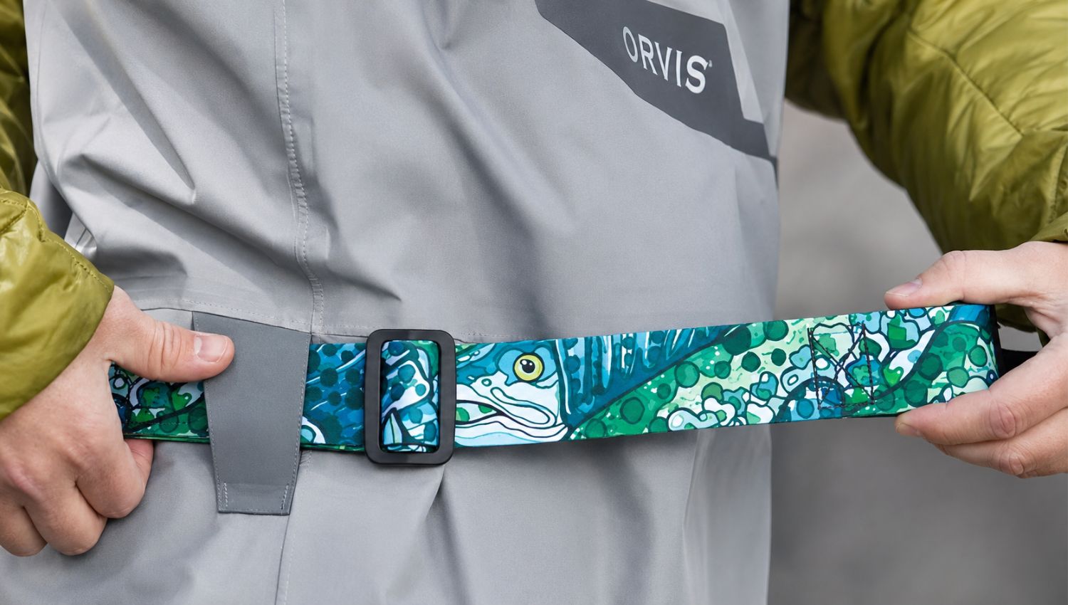 Person holding a colorful belt with a design on a gray Orvis jacket.