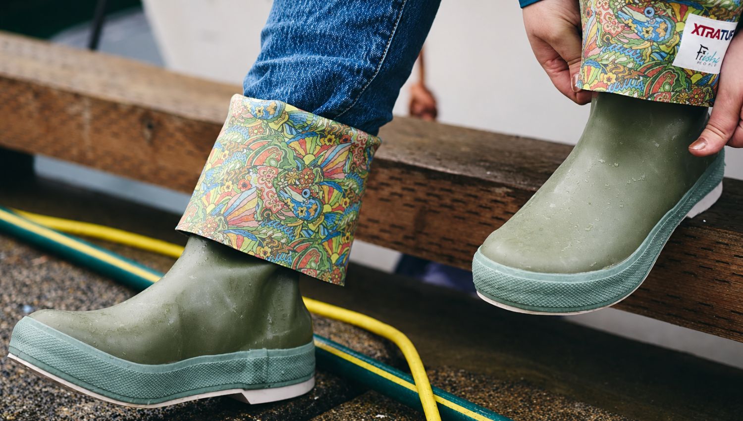 Green rubber boots with patterned cuffs worn by a person on a wooden bench.