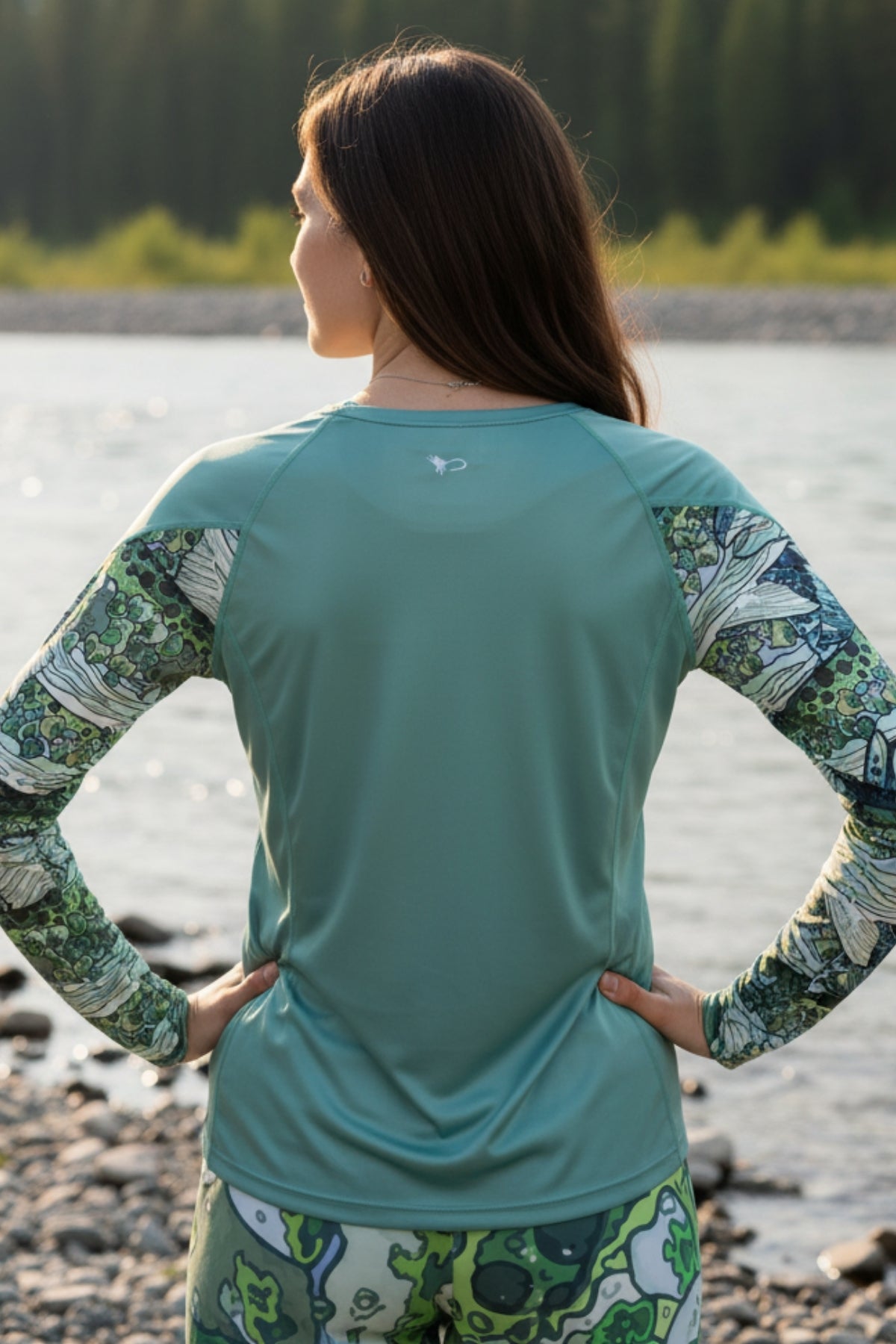 Person wearing a teal long-sleeve shirt with patterned sleeves and pants by a body of water.