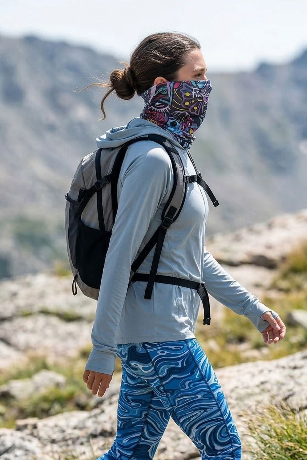 Person hiking in mountains wearing a colorful face mask, gray hoodie, and blue patterned pants.