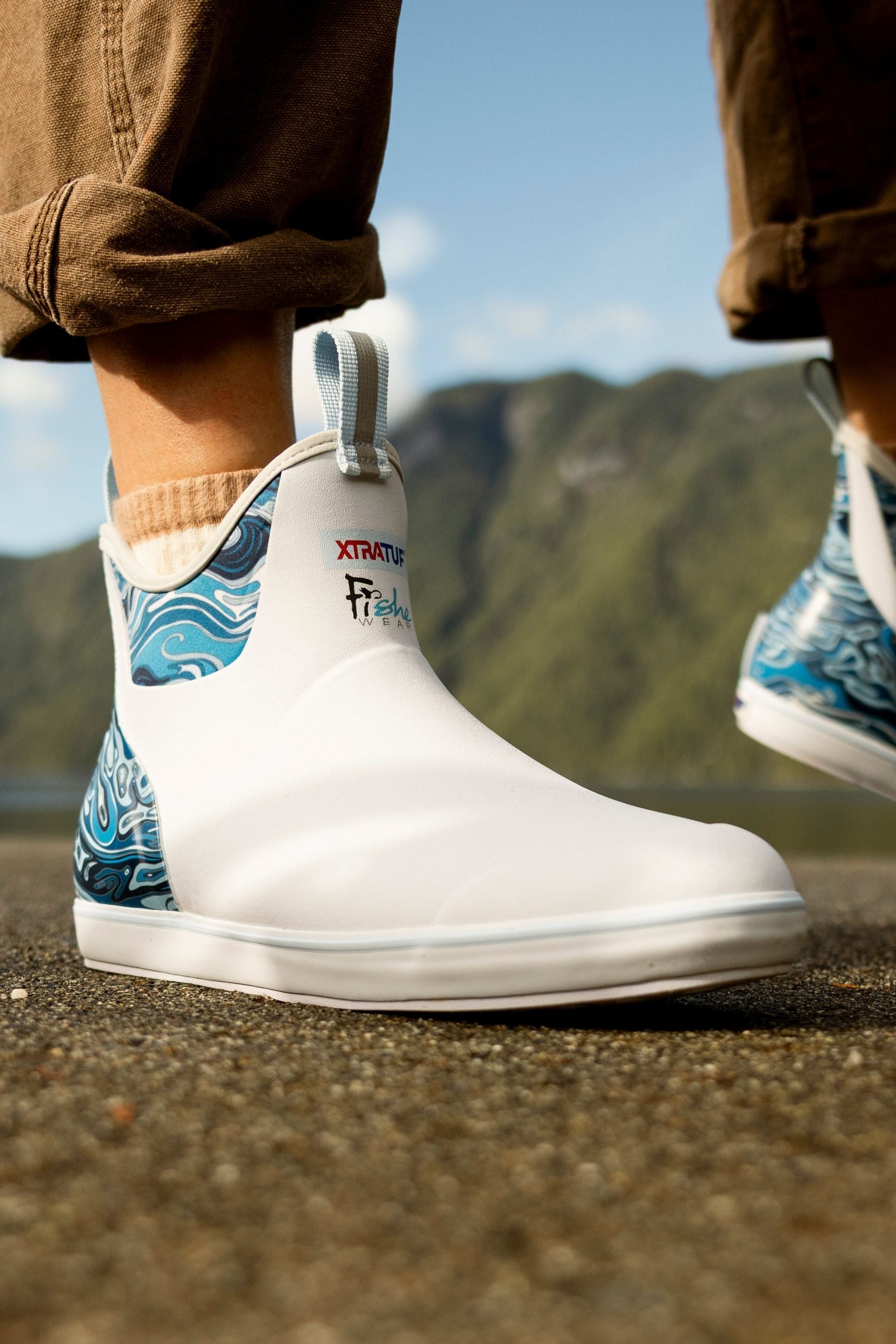 White rain boots with blue wave design worn by a person standing on a mountain path.