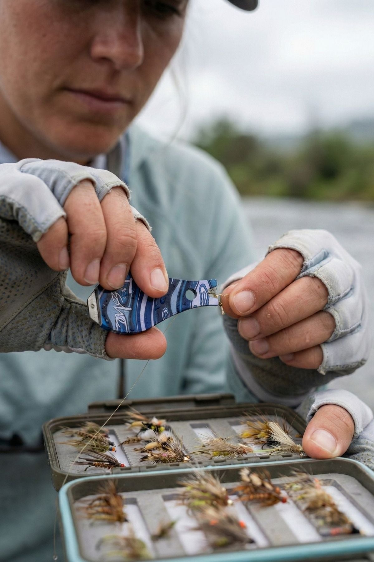 Person holding a fly fishing box with flies, wearing gloves and a cap.