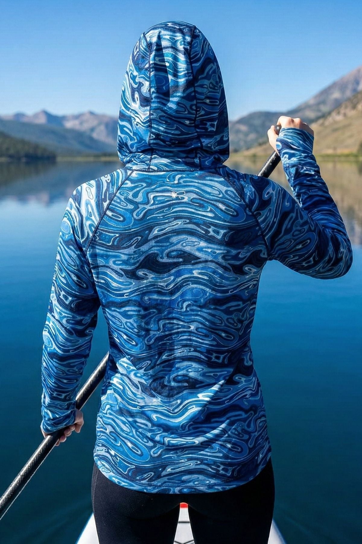 Person wearing a blue patterned hoodie standing by a lake with mountains in the background