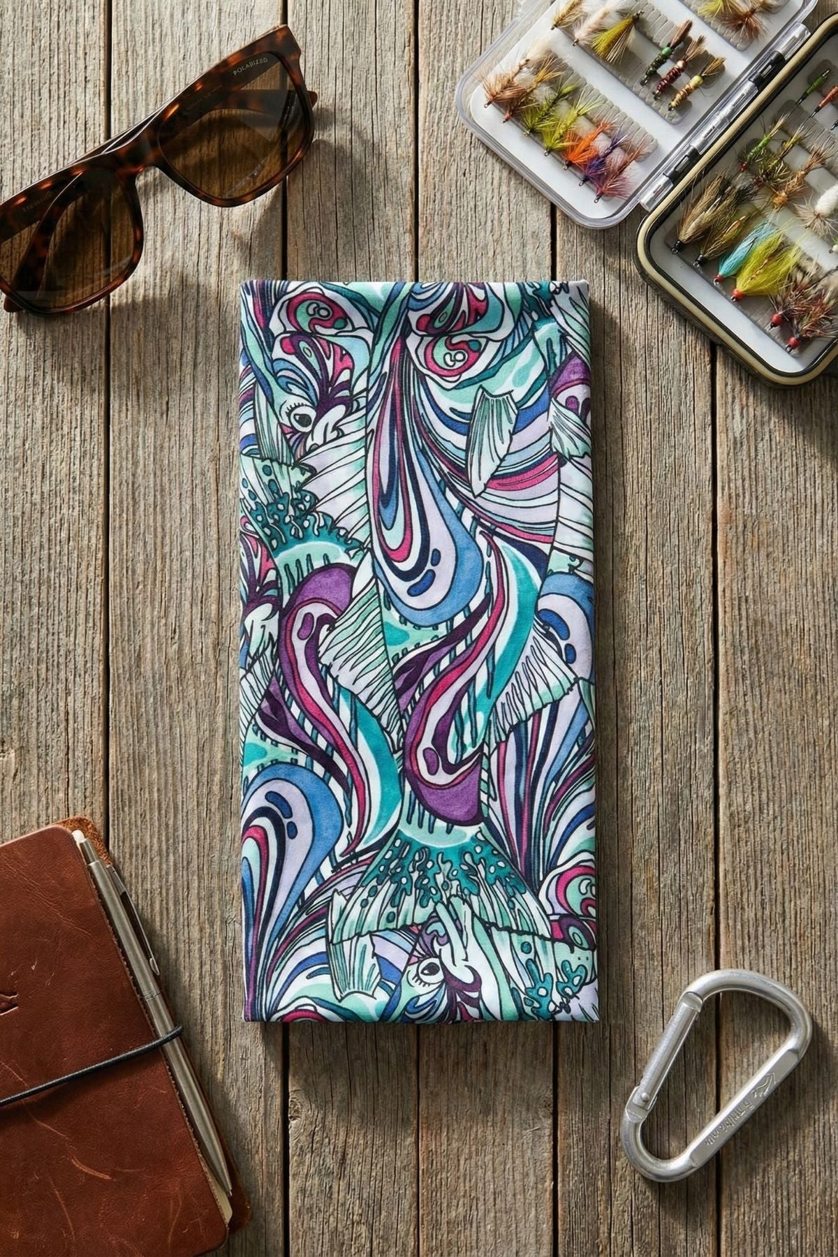 Colorful patterned phone case on a wooden surface with sunglasses, fly fishing tackle box, and carabiner.