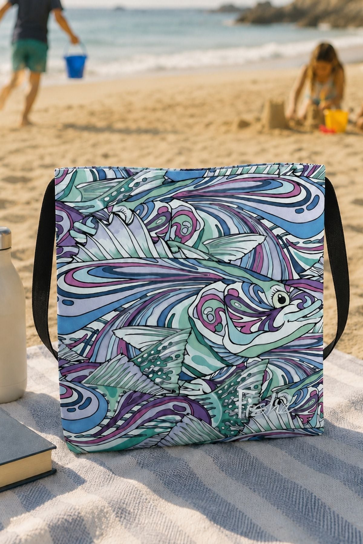 Colorful fish-patterned bag on a sandy beach with people in the background