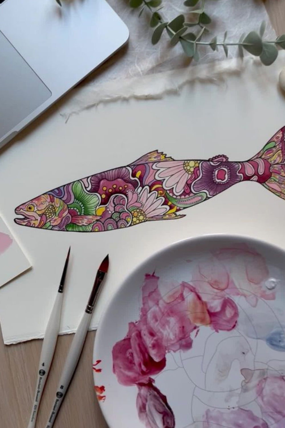 Colorful fish artwork beside paintbrushes and a paint palette.