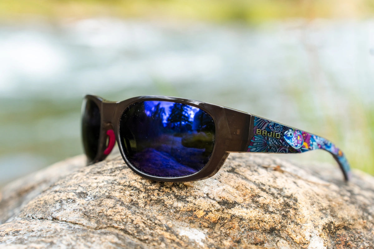 Bajío Darien sunglasses with blue mirrored lenses and Fishe Enchanted Grayling artwork beside the river